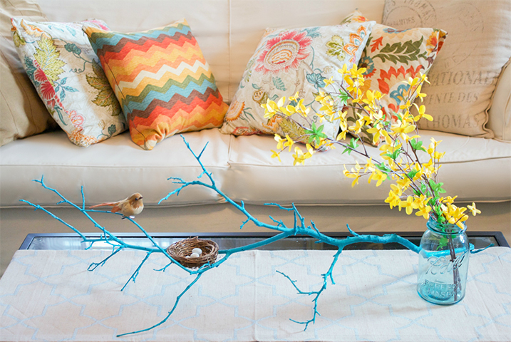 A coffee table with a blue branch holding a decorative bird and nest beside a small glass vase with yellow flowers in it.